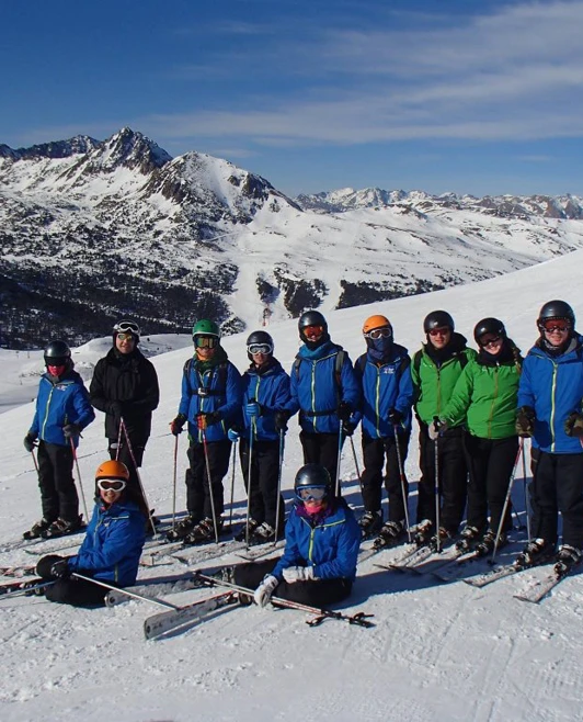 A group of students and staff pose with skis on a snowy mountain slope under clear blue skies. A group of students and staff pose with skis on a snowy mountain slope under clear blue skies.